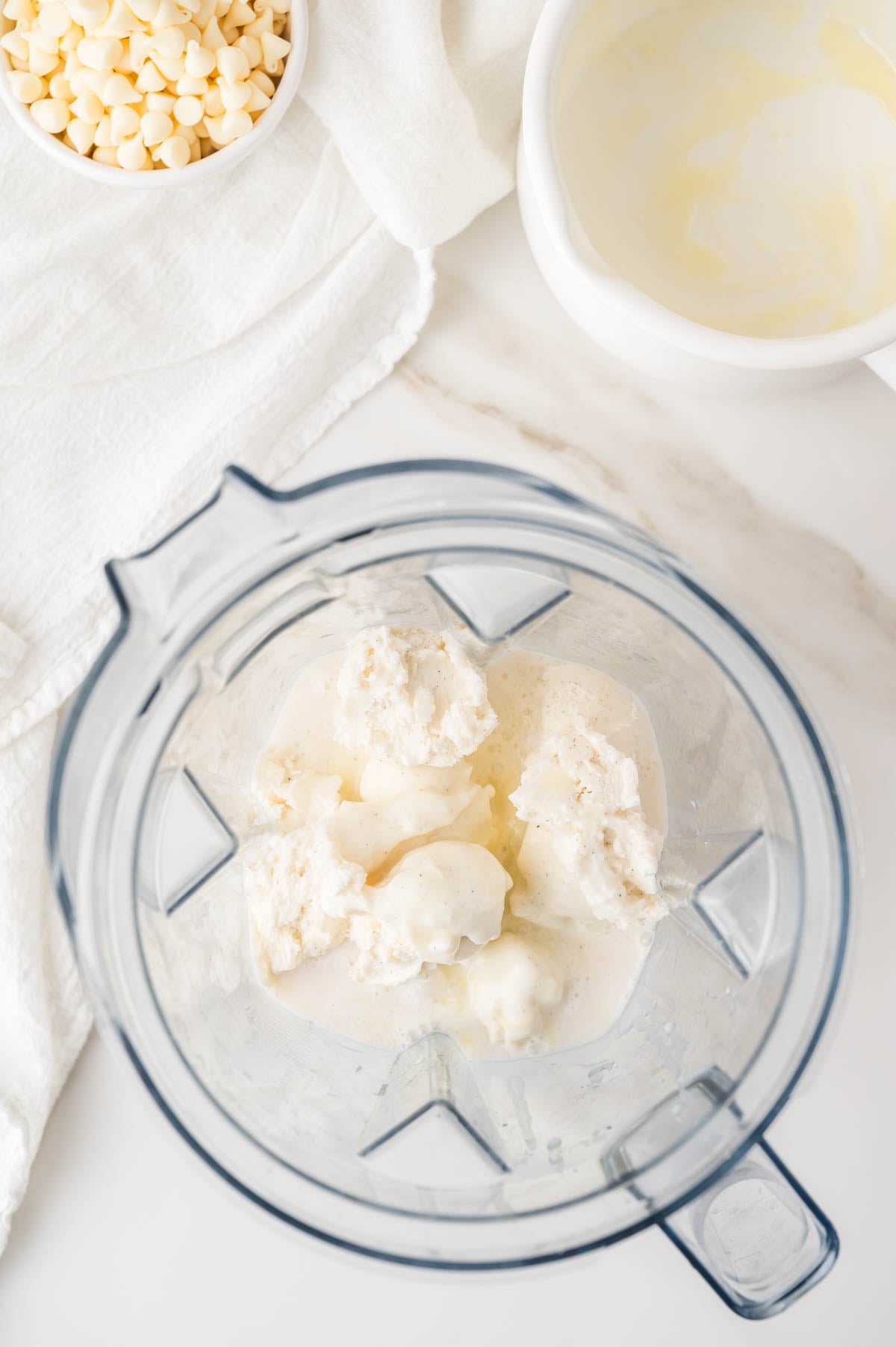 A blender contains scoops of vanilla ice cream and milk, with a white cloth, a bowl of white chocolate chips, and an empty bowl nearby on a white surface.