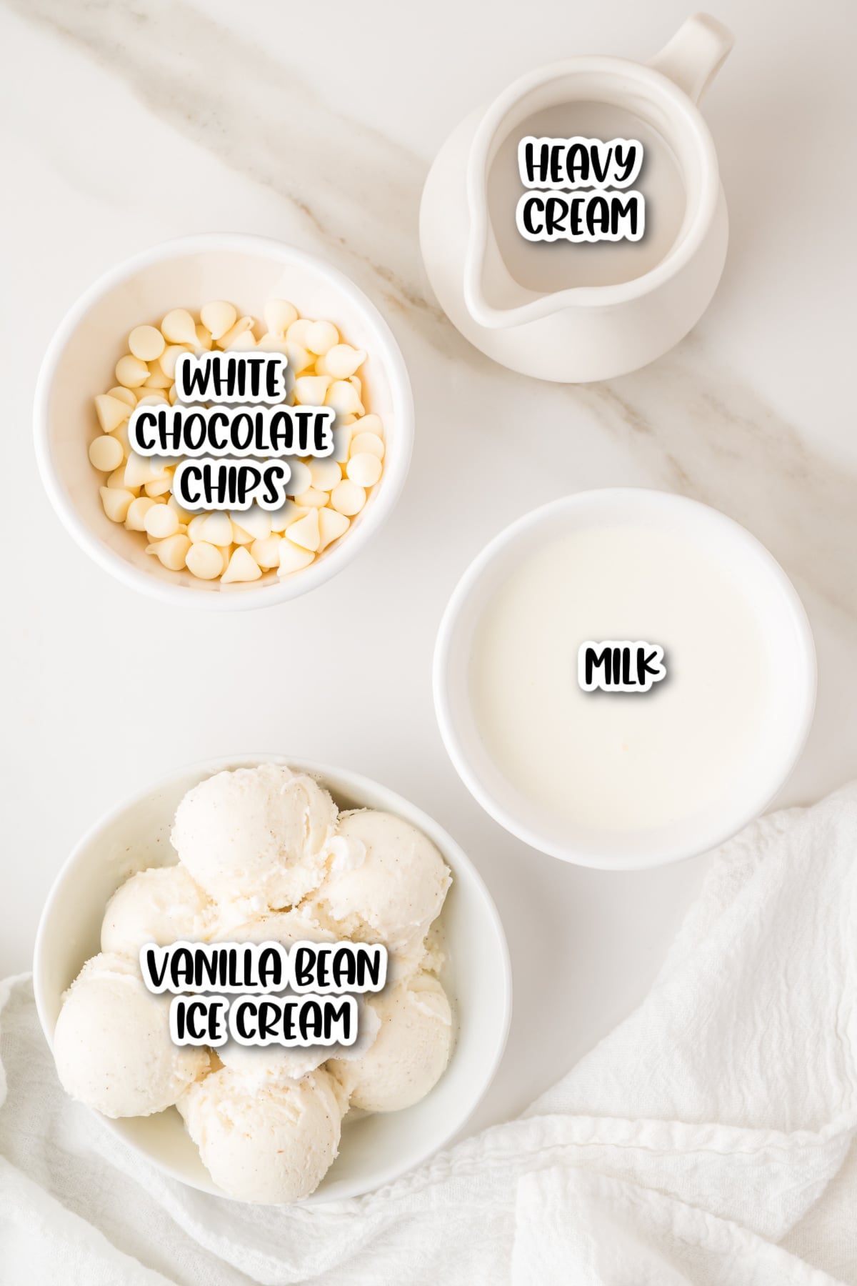 Bowls of white chocolate chips, vanilla bean ice cream, and milk, with a small pitcher of heavy cream on a white surface. Each item is labeled.