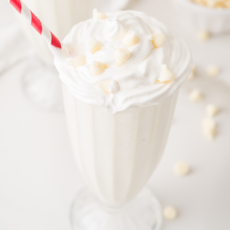 A vanilla milkshake topped with whipped cream and white chocolate chips in a glass, with a red and white striped straw.