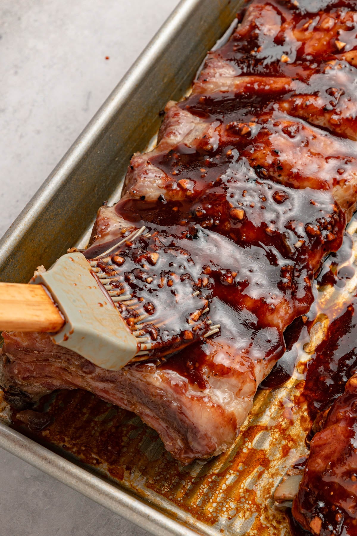 A rack of ribs in a metal baking pan being brushed with a thick, dark barbecue sauce using a silicone brush.