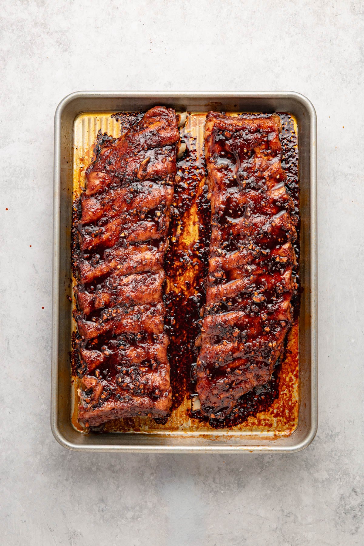 Two racks of glazed, cooked ribs on a rectangular metal baking tray with charred edges, viewed from above on a light-colored surface.