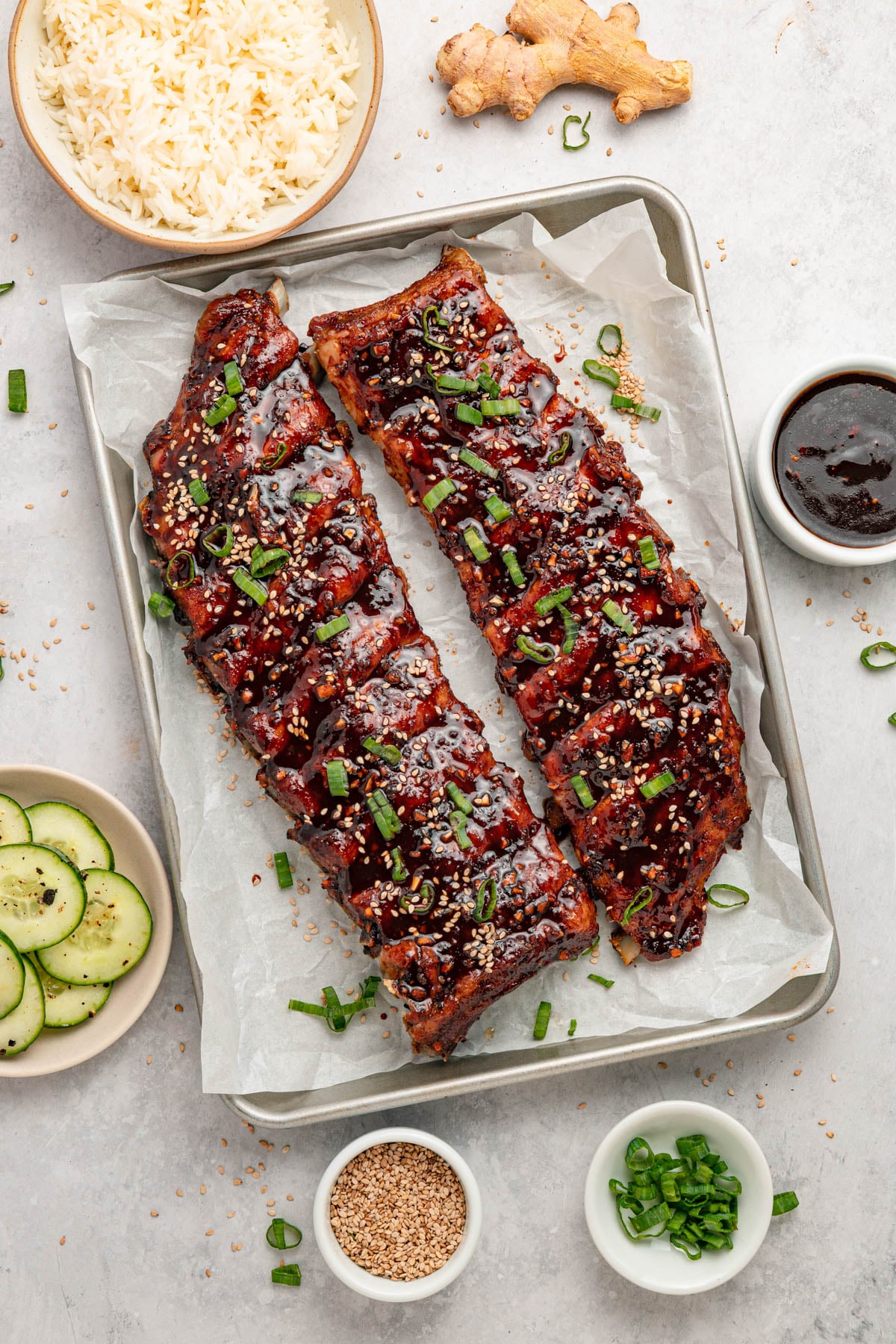 Two racks of glazed ribs garnished with sesame seeds and green onions on a baking sheet, surrounded by bowls of rice, cucumber slices, sauce, sesame seeds, and chopped green onions.
