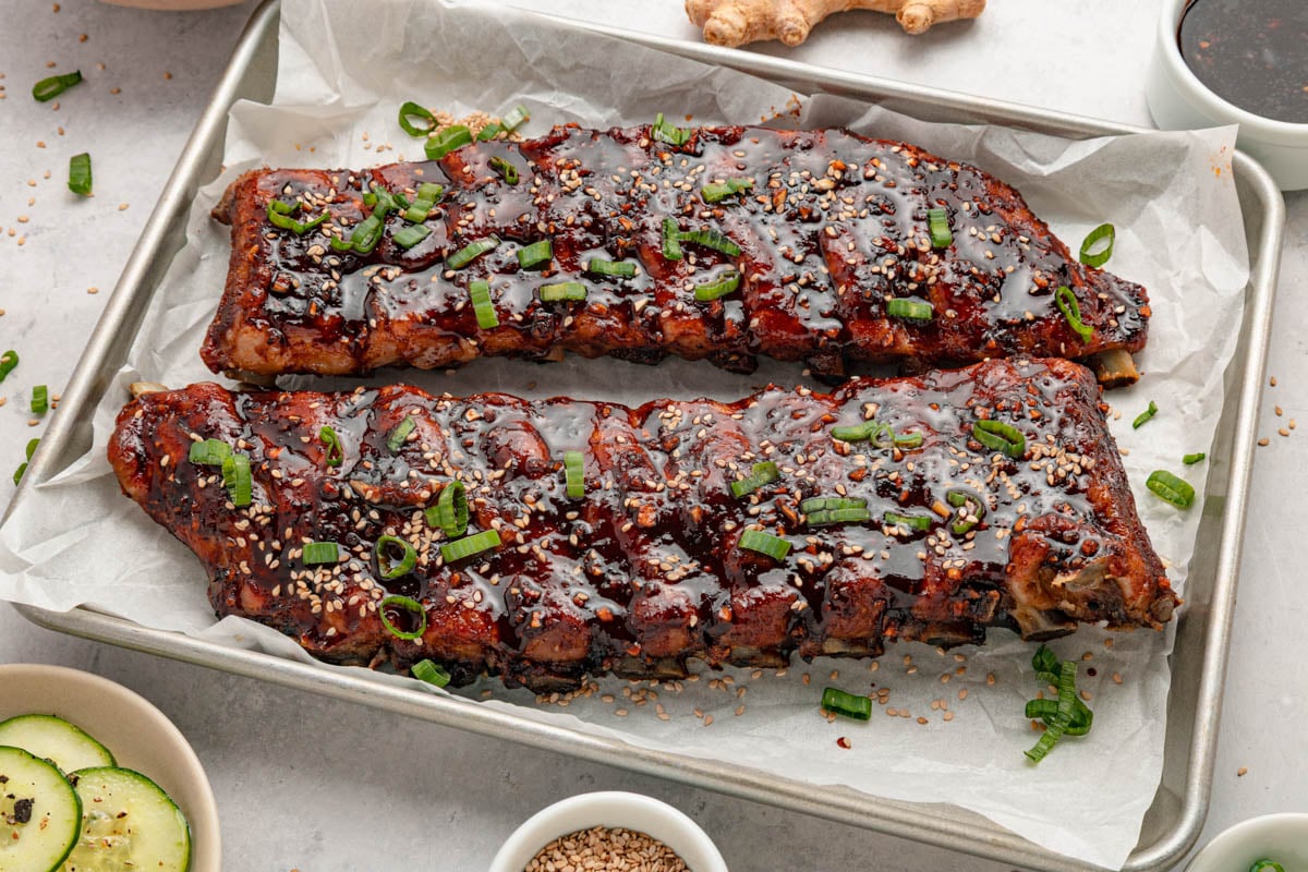 Two racks of glazed ribs garnished with chopped green onions and sesame seeds on a parchment-lined baking tray, with side dishes nearby.