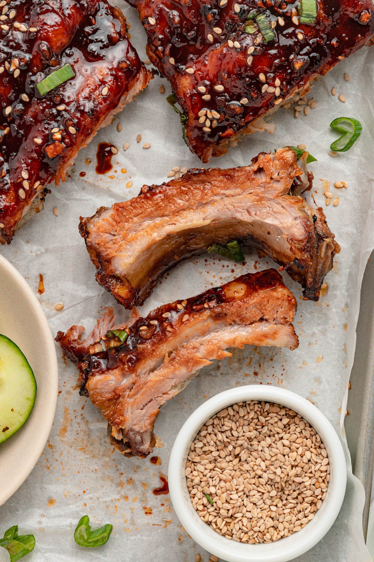 Cooked pork ribs with a glossy sauce, garnished with sesame seeds and green onions, are arranged on parchment paper next to a small bowl of sesame seeds.