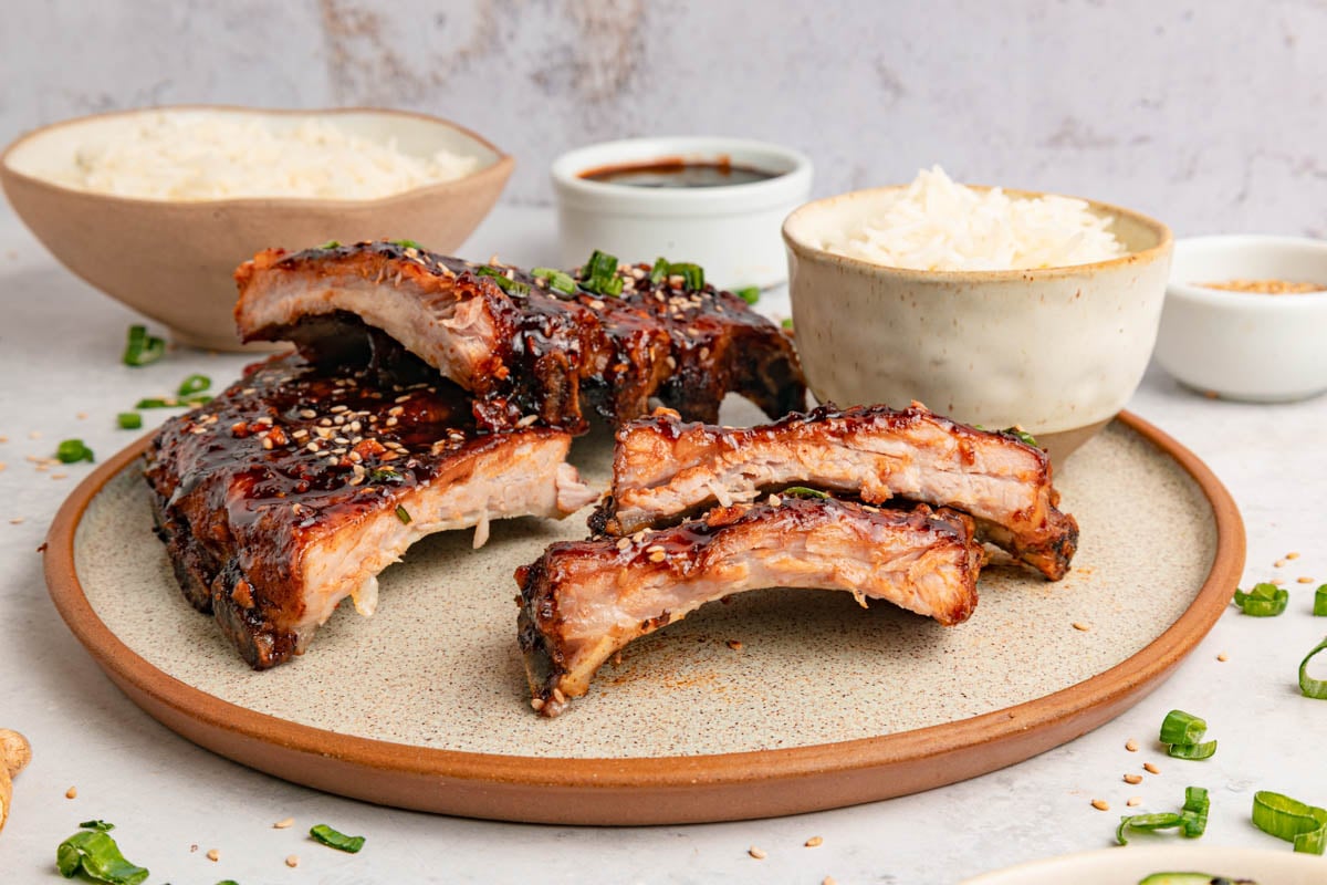 A plate of glazed pork ribs garnished with sesame seeds and green onions, served with bowls of white rice and sauce on a light tabletop.
