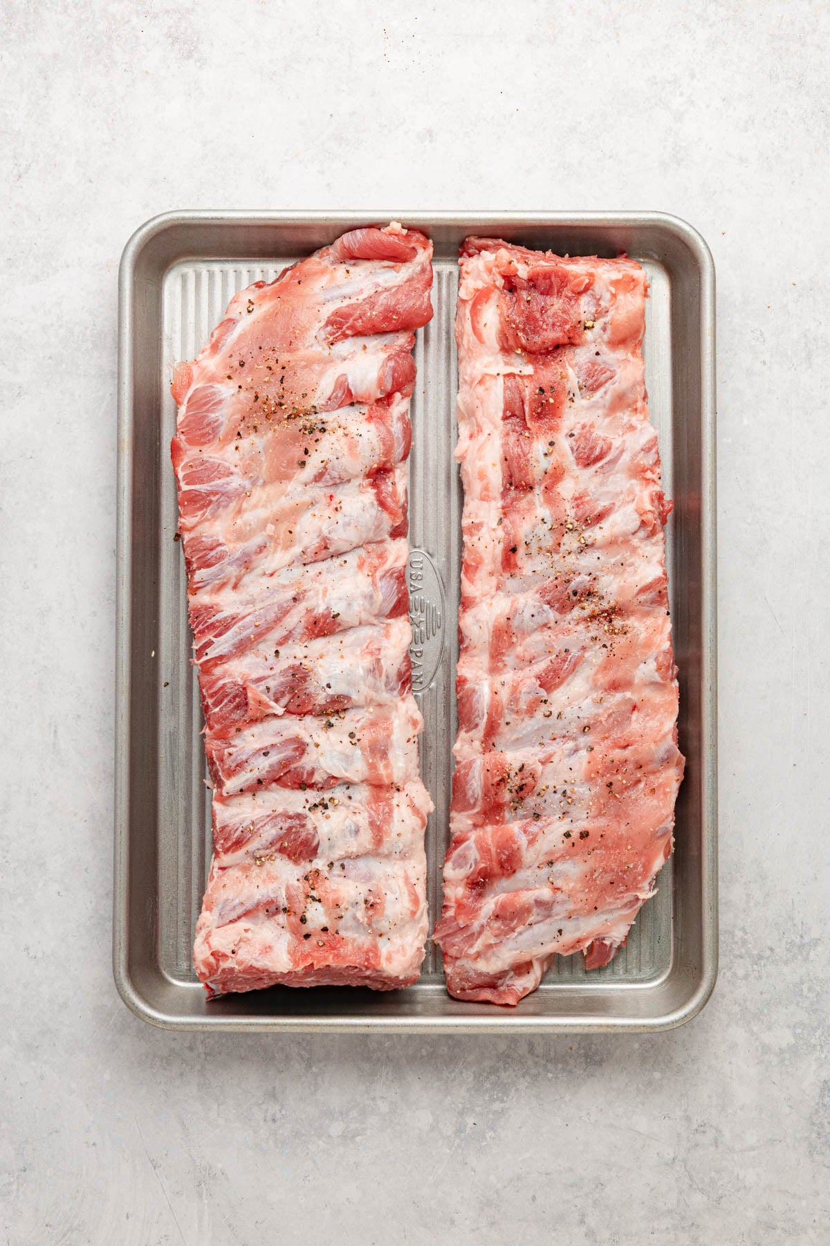 Two raw racks of pork ribs with seasoning are placed side by side on a metal baking tray, viewed from above on a light-colored surface.
