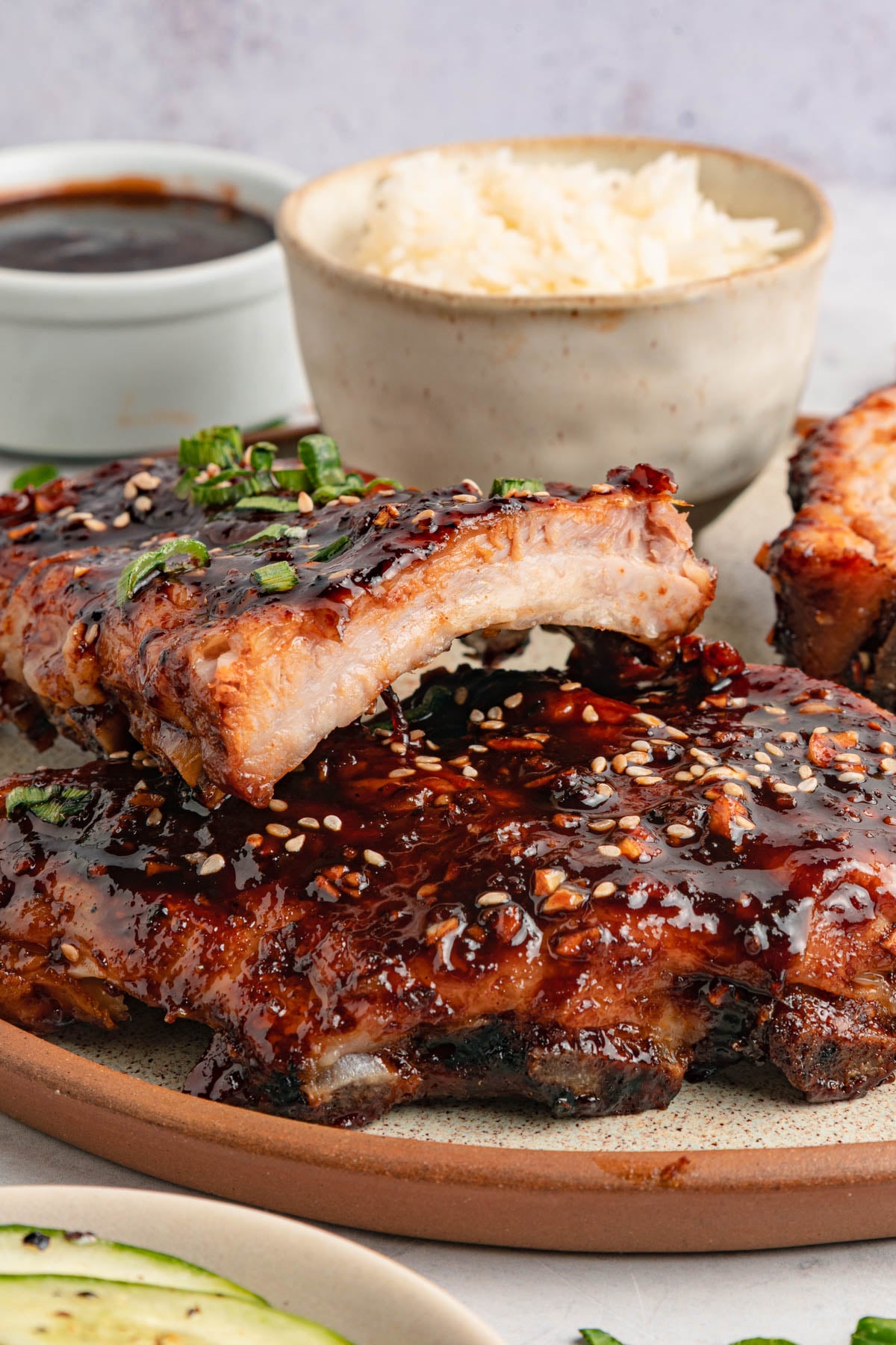 A plate of glazed ribs garnished with sesame seeds and green onions, with a bowl of rice and a dish of sauce in the background.