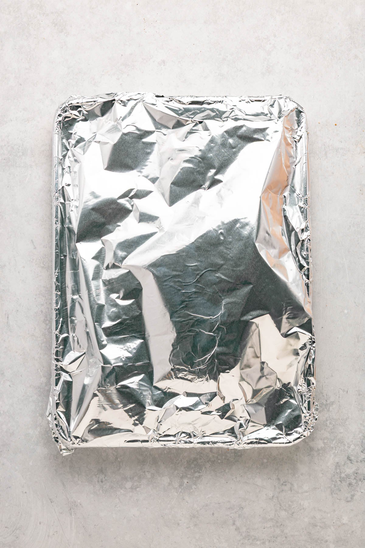 A rectangular baking dish covered tightly with aluminum foil is placed on a light gray surface.