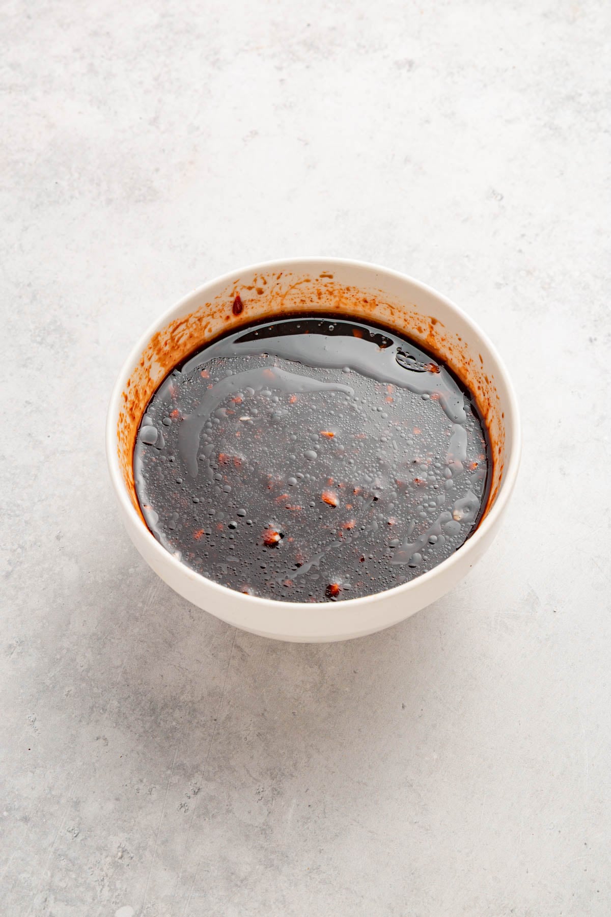 A white bowl filled with a dark brown liquid sauce with small bits, placed on a light gray surface.
