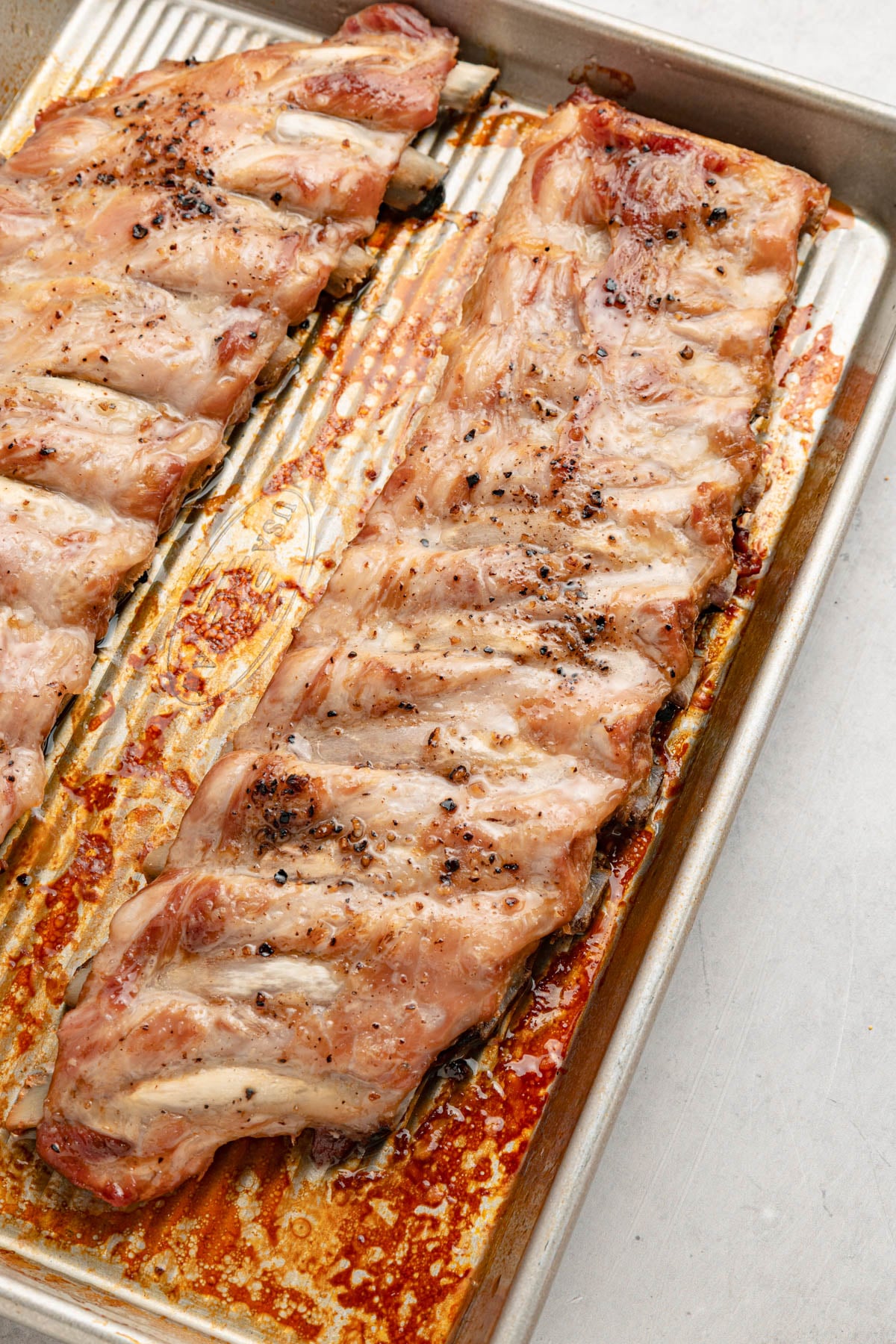 Two racks of cooked pork ribs seasoned with pepper on a metal baking sheet, with juices and browned spots visible.