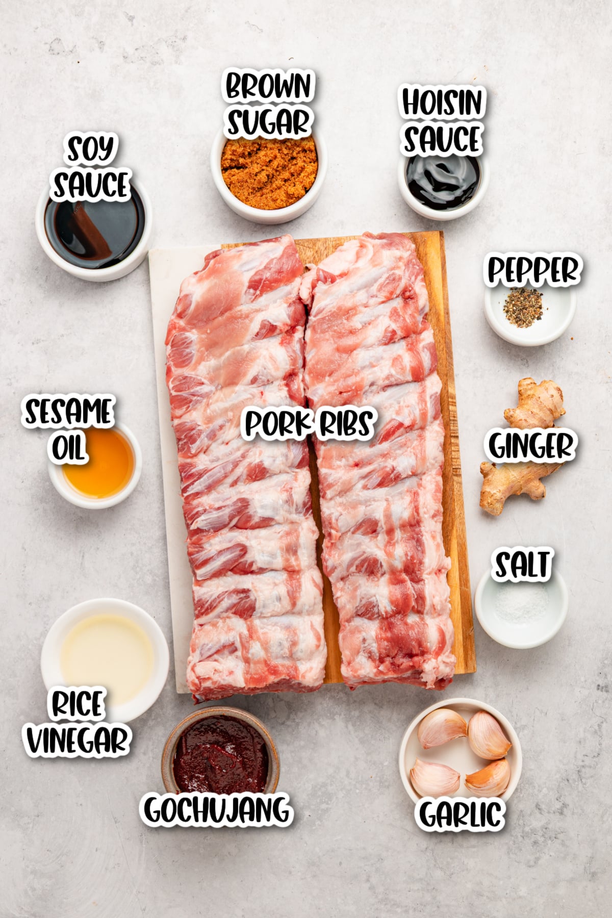 Two racks of raw pork ribs on a cutting board, surrounded by small bowls of soy sauce, sesame oil, rice vinegar, brown sugar, gochujang, hoisin sauce, salt, pepper, ginger, and garlic.