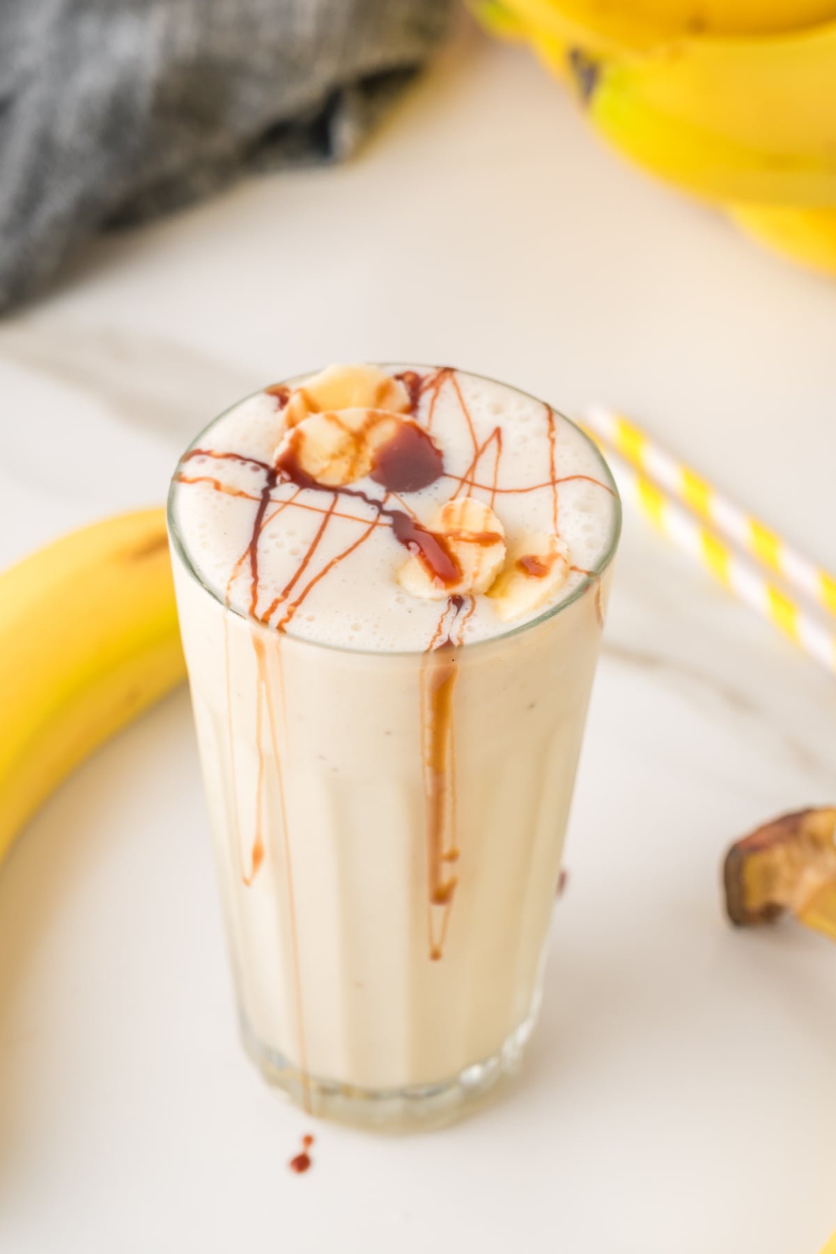 A glass of banana smoothie topped with banana slices and chocolate syrup, with a yellow-striped straw and bananas in the background.