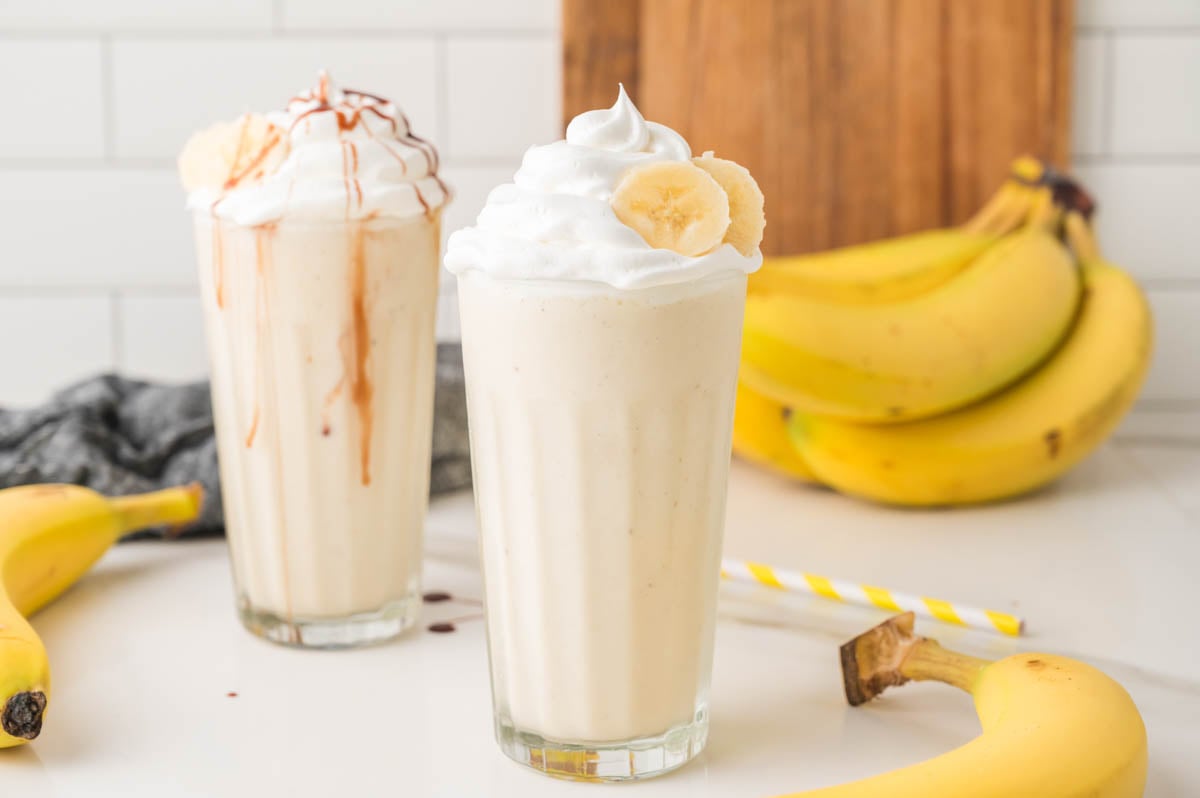 Two banana milkshakes in tall glasses, one topped with whipped cream and banana slices, the other with whipped cream and caramel drizzle, with bananas and straws in the background.