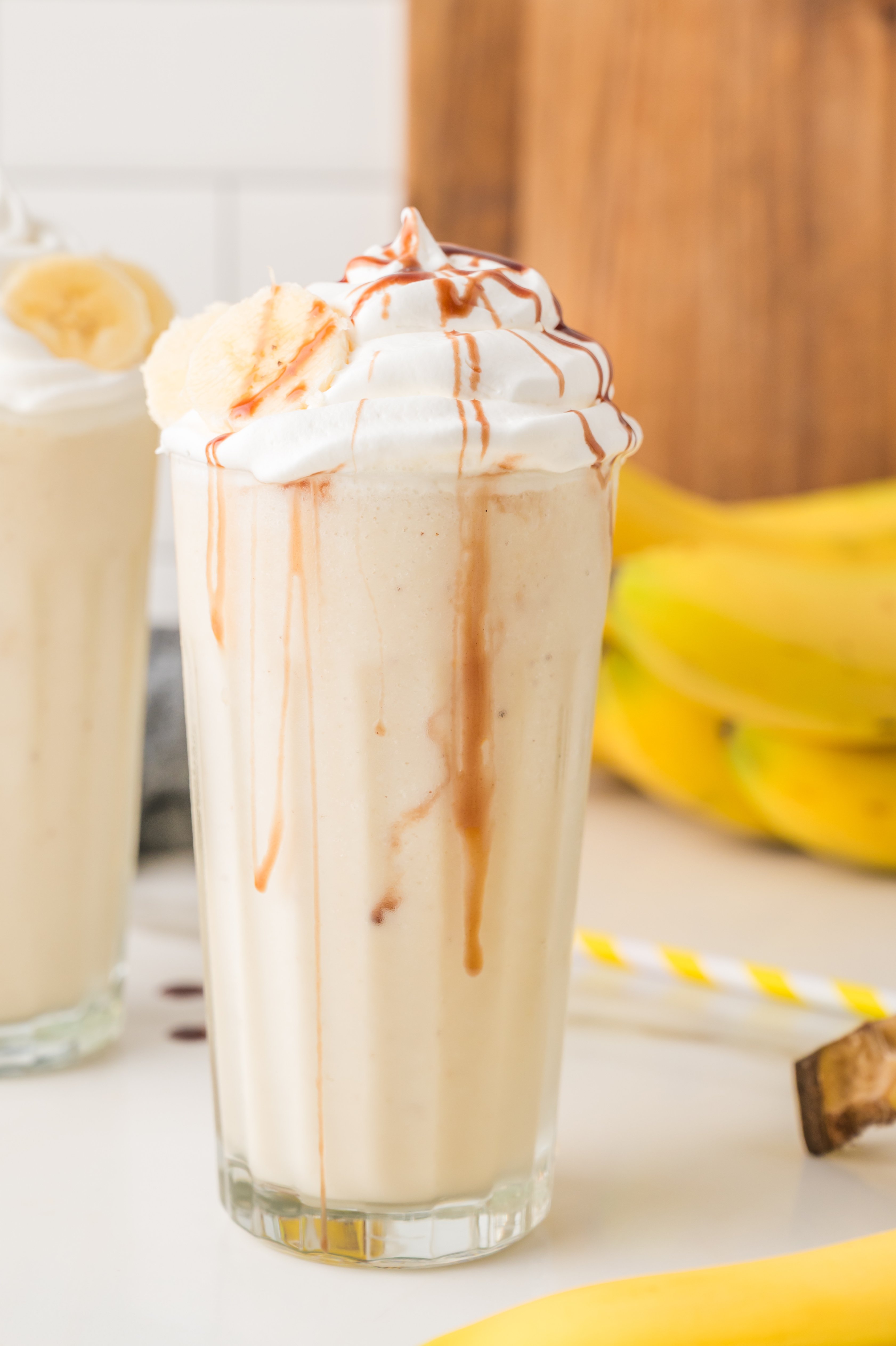 A tall glass of banana milkshake topped with whipped cream and chocolate drizzle, with banana slices and whole bananas in the background.