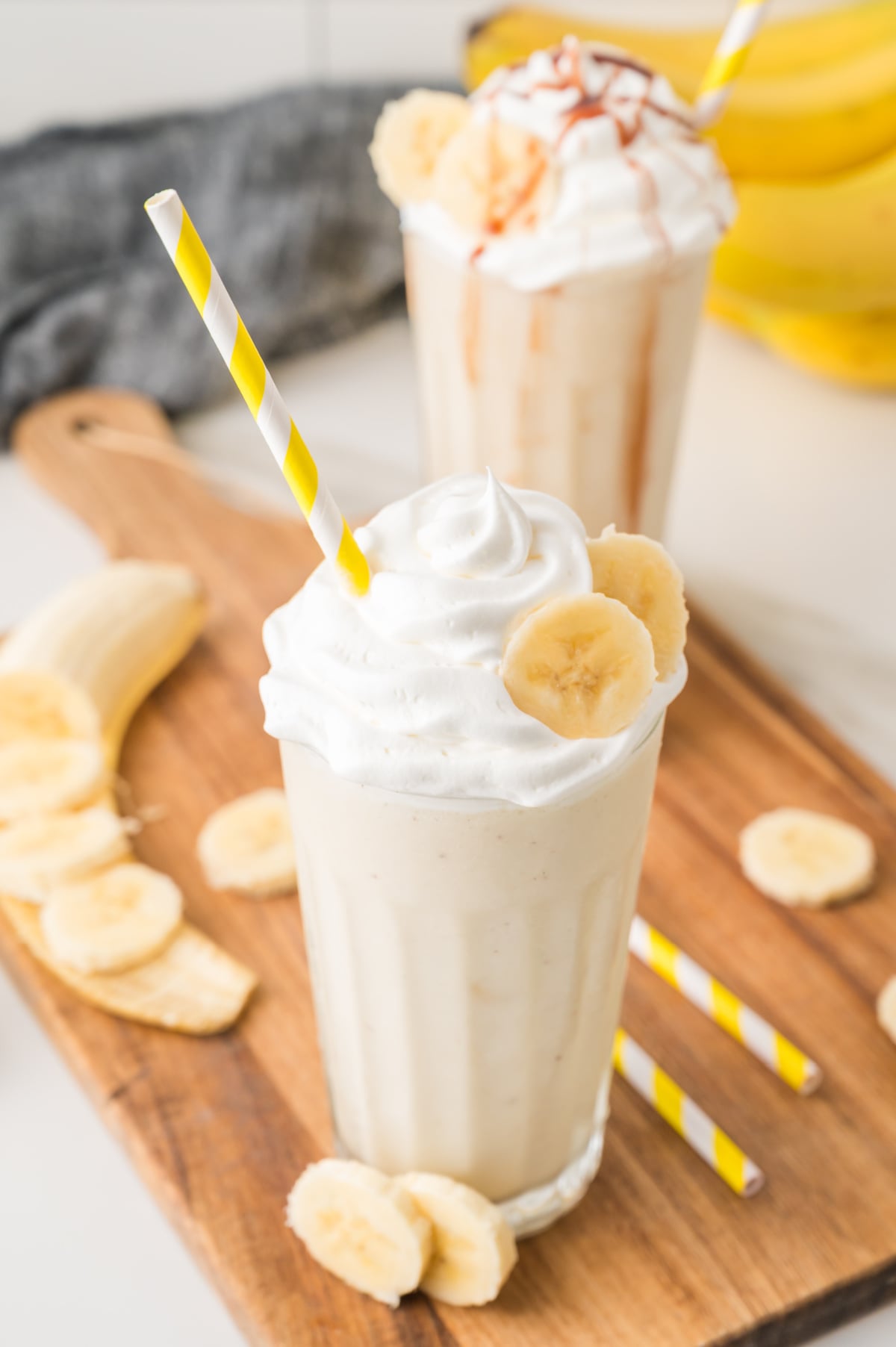 Two banana milkshakes with whipped cream and banana slices, each with a yellow-striped straw, on a wooden board with scattered banana pieces.