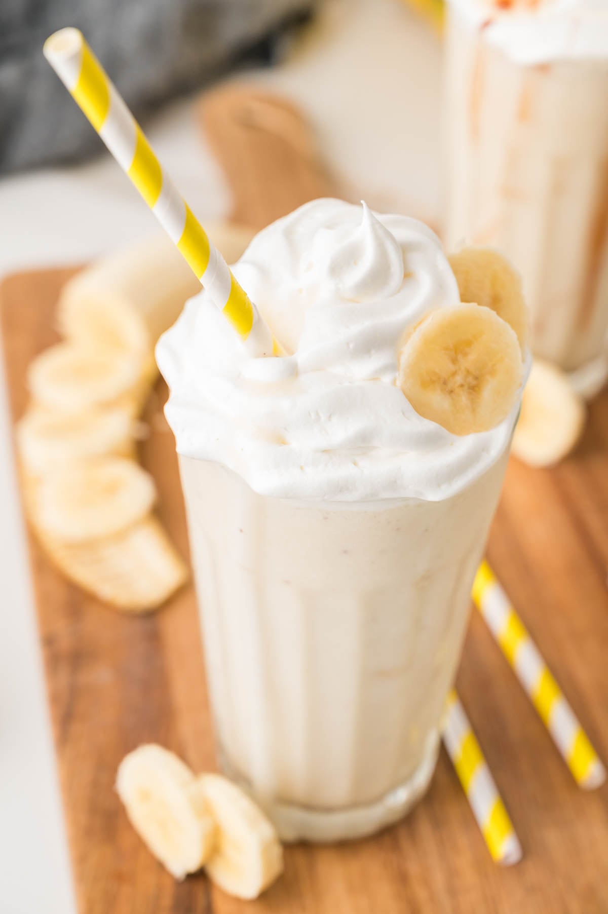 A glass of banana milkshake topped with whipped cream, garnished with banana slices, and served with a yellow-striped straw on a wooden surface.