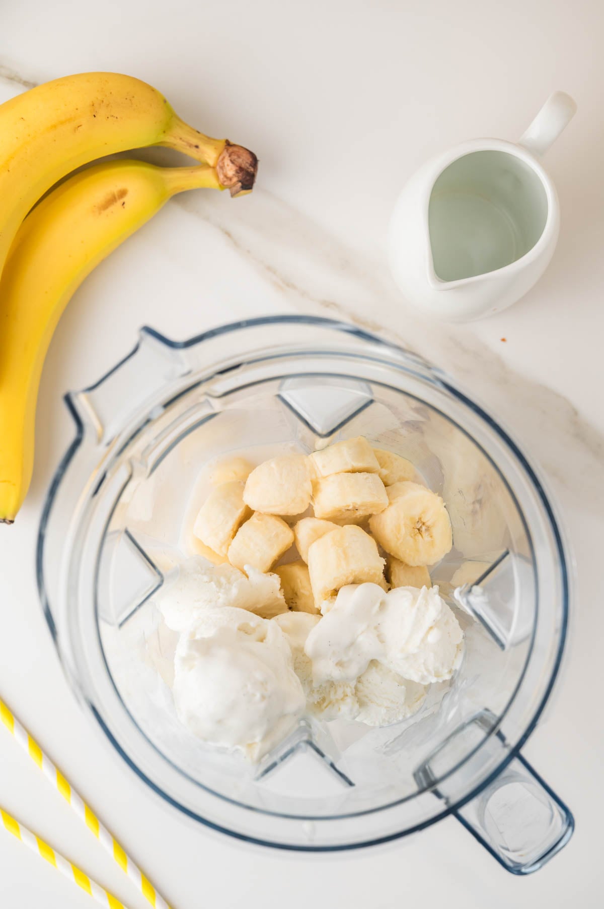 A blender contains sliced bananas and scoops of vanilla ice cream, with two bananas, a small pitcher of milk, and two yellow-striped straws on the counter nearby.