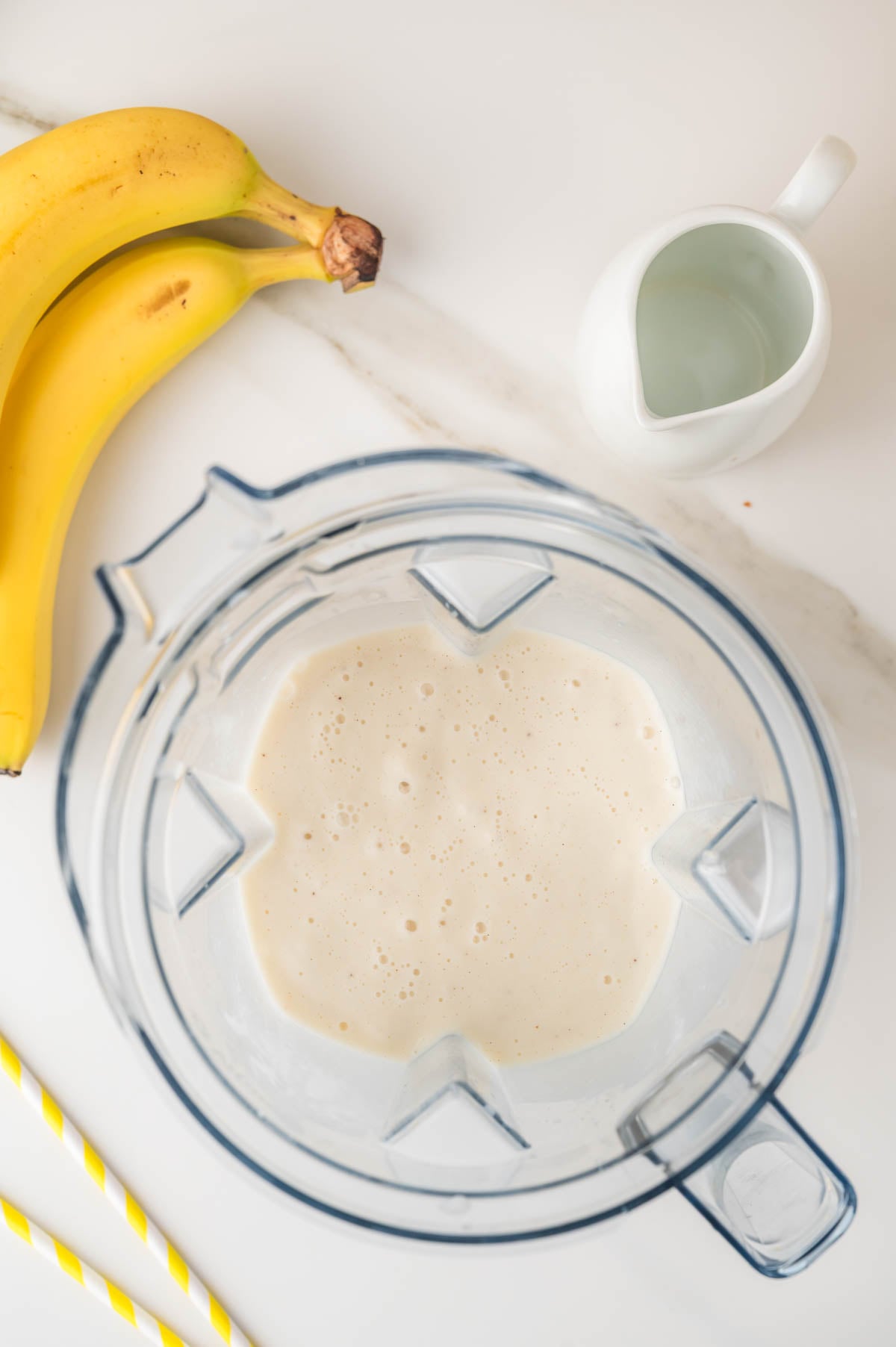 Overhead view of a blender containing a creamy, blended mixture, with two bananas, a small pitcher of liquid, and two yellow-striped straws nearby on a white surface.