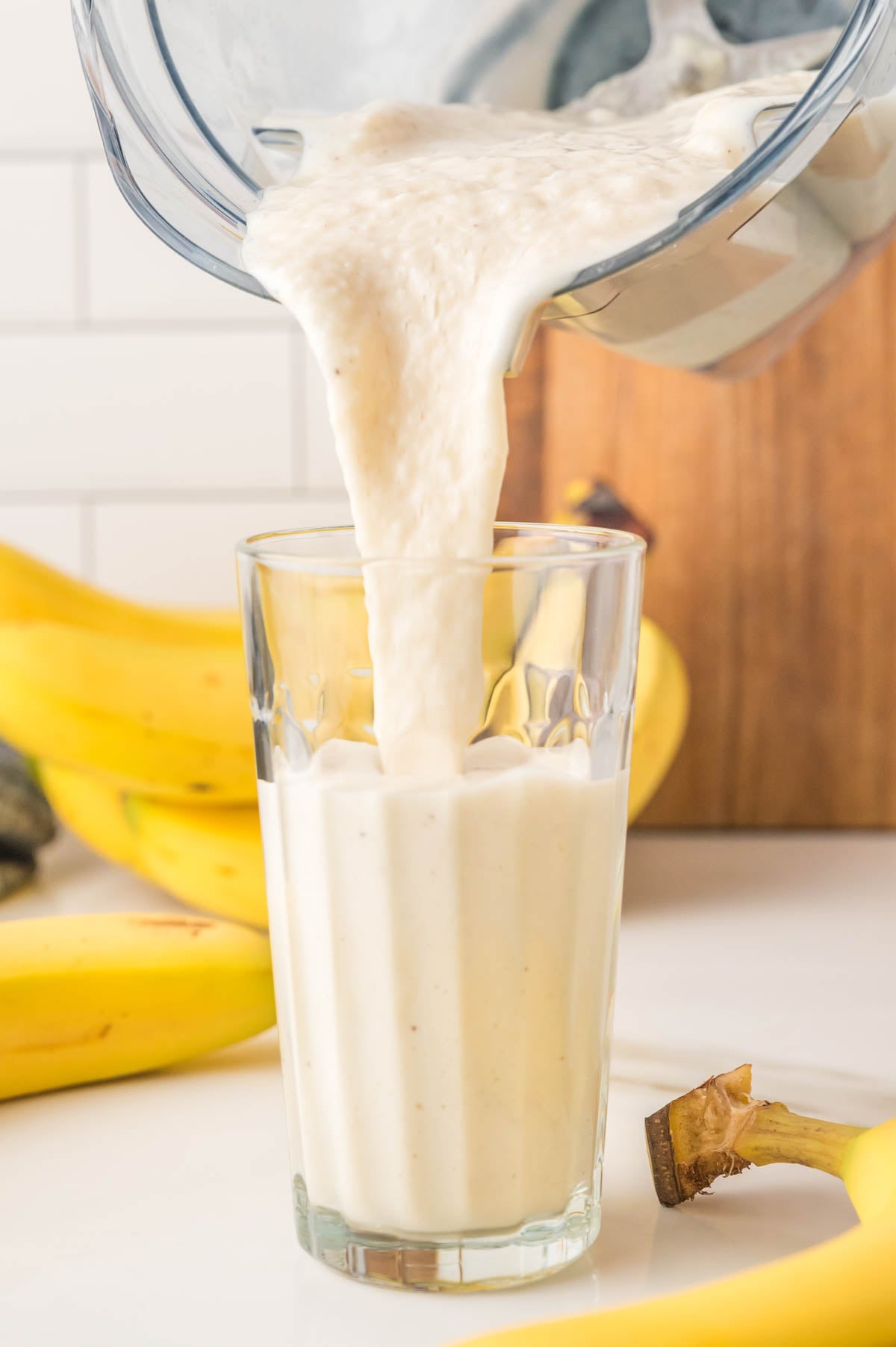 A thick banana smoothie is being poured from a blender into a glass, with ripe bananas visible in the background.