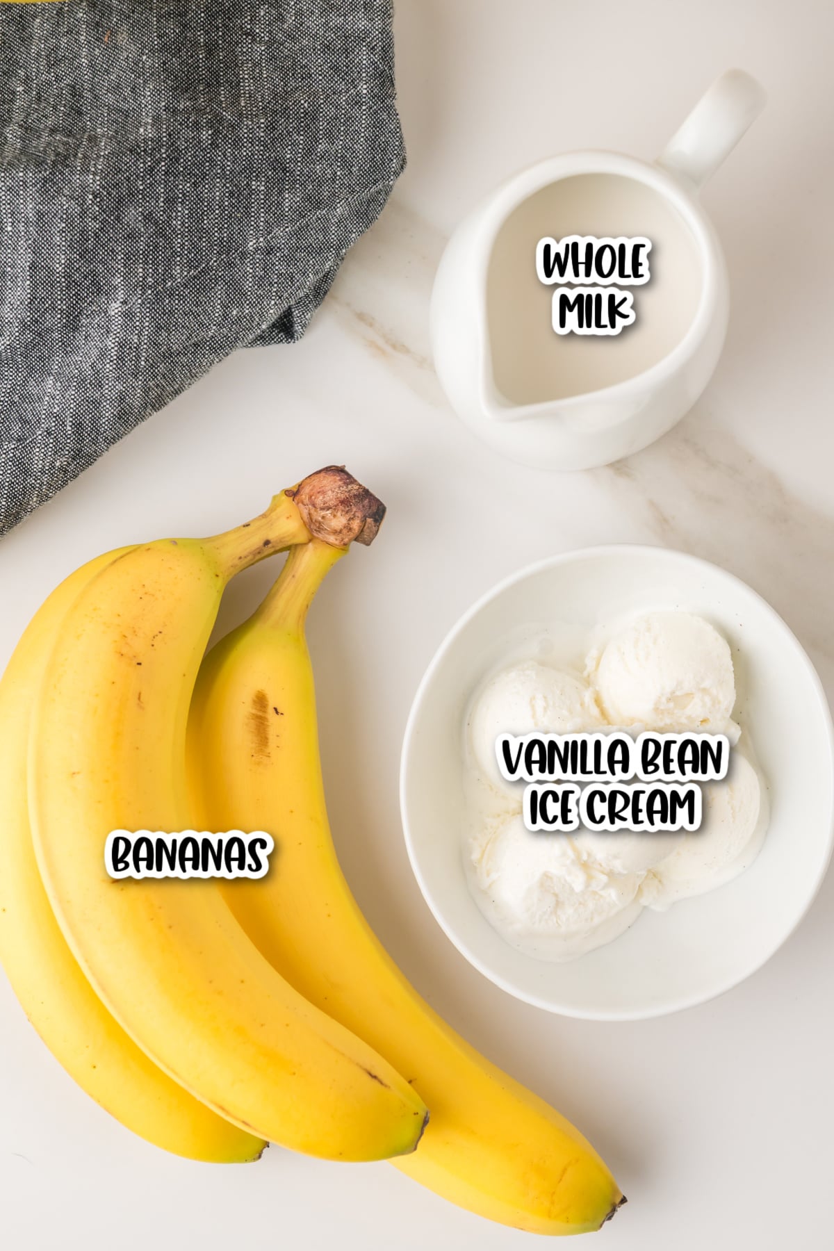 A bunch of bananas, a bowl of vanilla bean ice cream, and a small pitcher of whole milk arranged on a white surface.