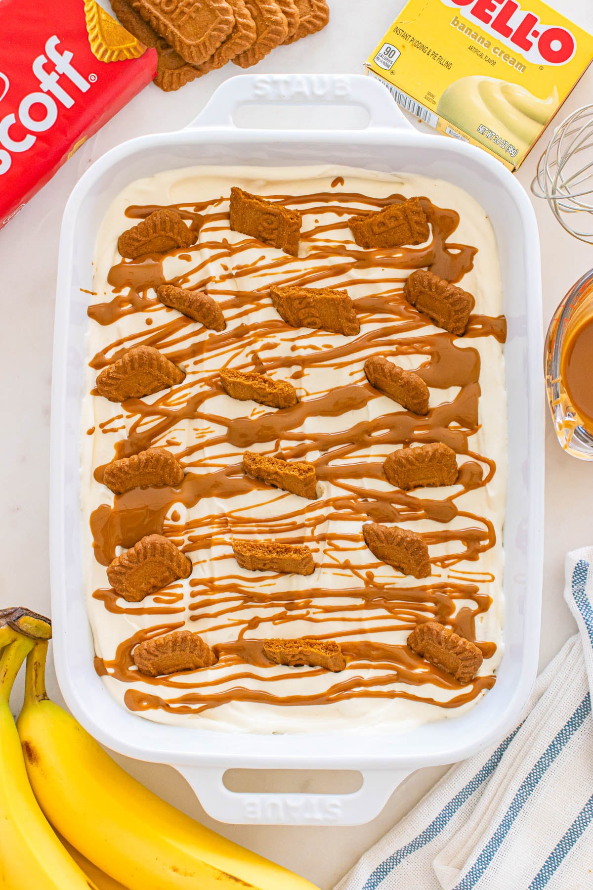 A white baking dish filled with banana cream pudding, topped with Biscoff cookies and caramel drizzle.