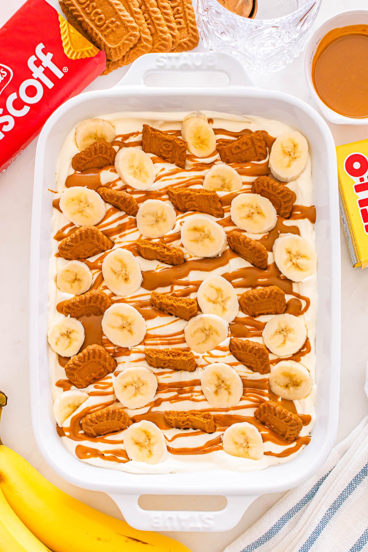 A white baking dish filled with a creamy dessert topped with banana slices, Biscoff cookies, and caramel drizzle.