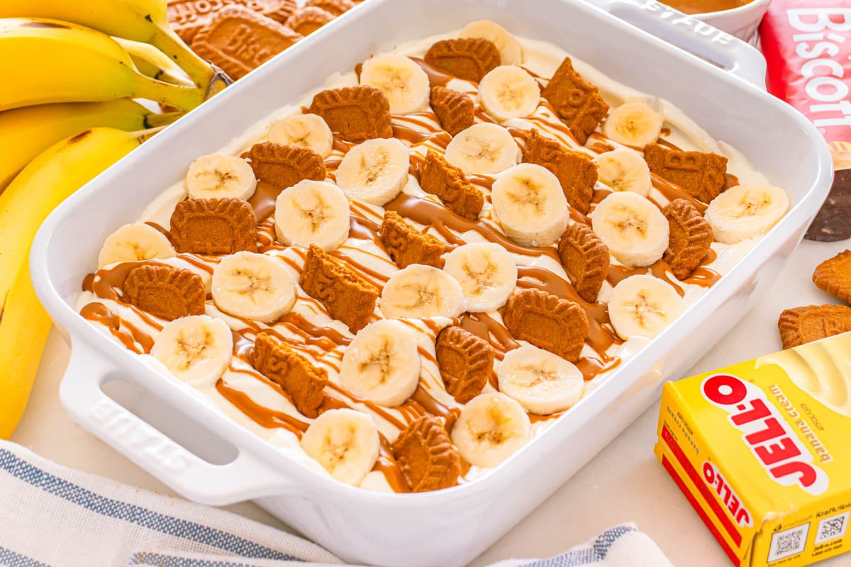 A white dish filled with banana pudding, topped with banana slices and speculoos cookies, drizzled with caramel sauce.