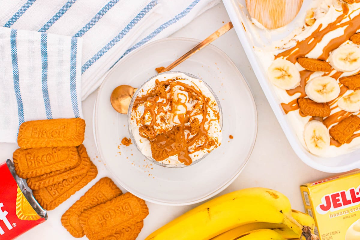 A glass filled with Biscoff Banana Pudding topped with crushed cookies sits on a plate, with Biscoff cookies, bananas, and a box of Jell-O banana cream pudding nearby.