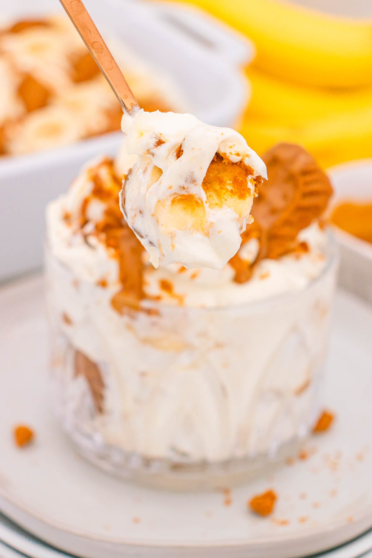 A spoon scoops a portion of banana pudding layered with cookies.from a glass dish, with bananas in the background.