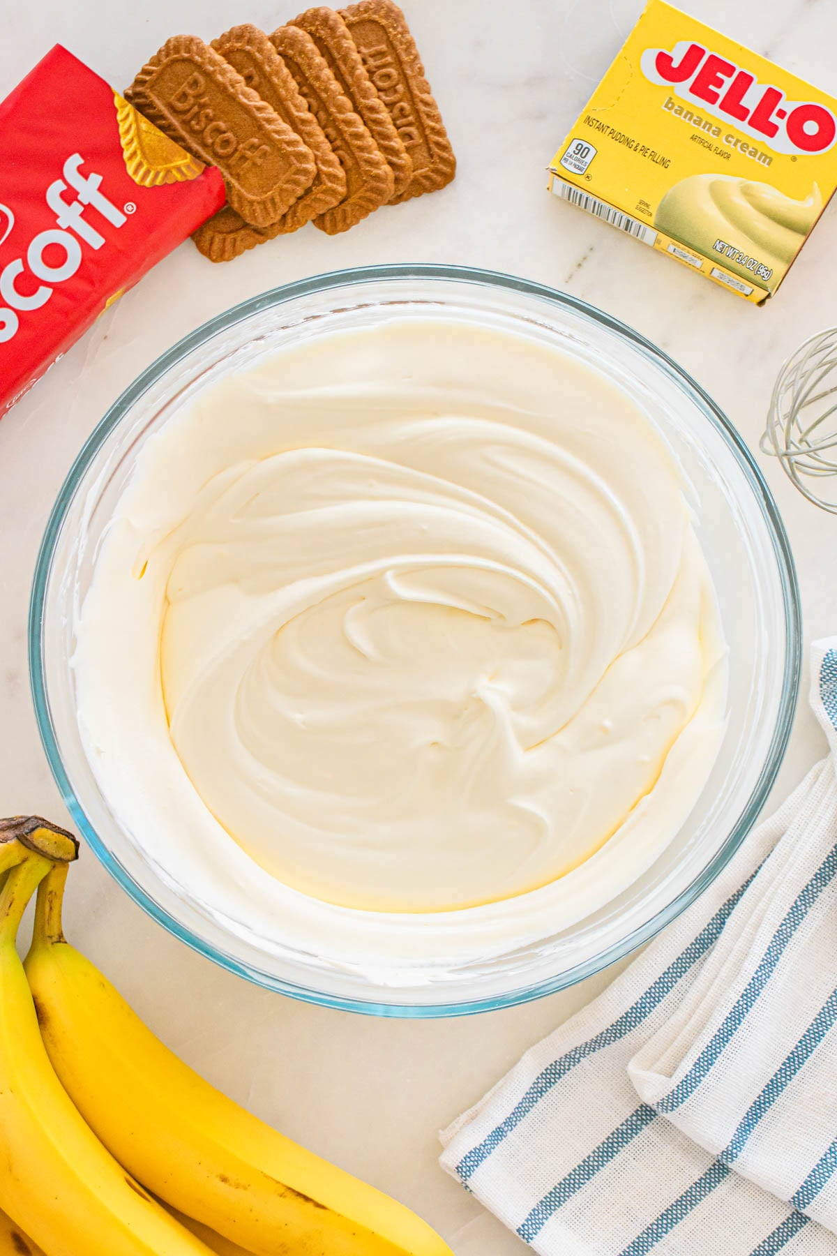 A bowl of smooth, creamy banana pudding and Cool Whip mixture is surrounded by Biscoff cookies, a box of Jell-O banana cream pudding, and two bananas.