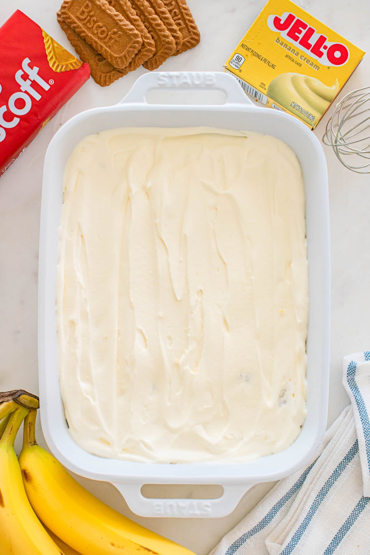A white baking dish filled with creamy banana pudding is surrounded by Biscoff cookies, bananas, a box of Jell-O banana cream pudding mix, and a whisk.
