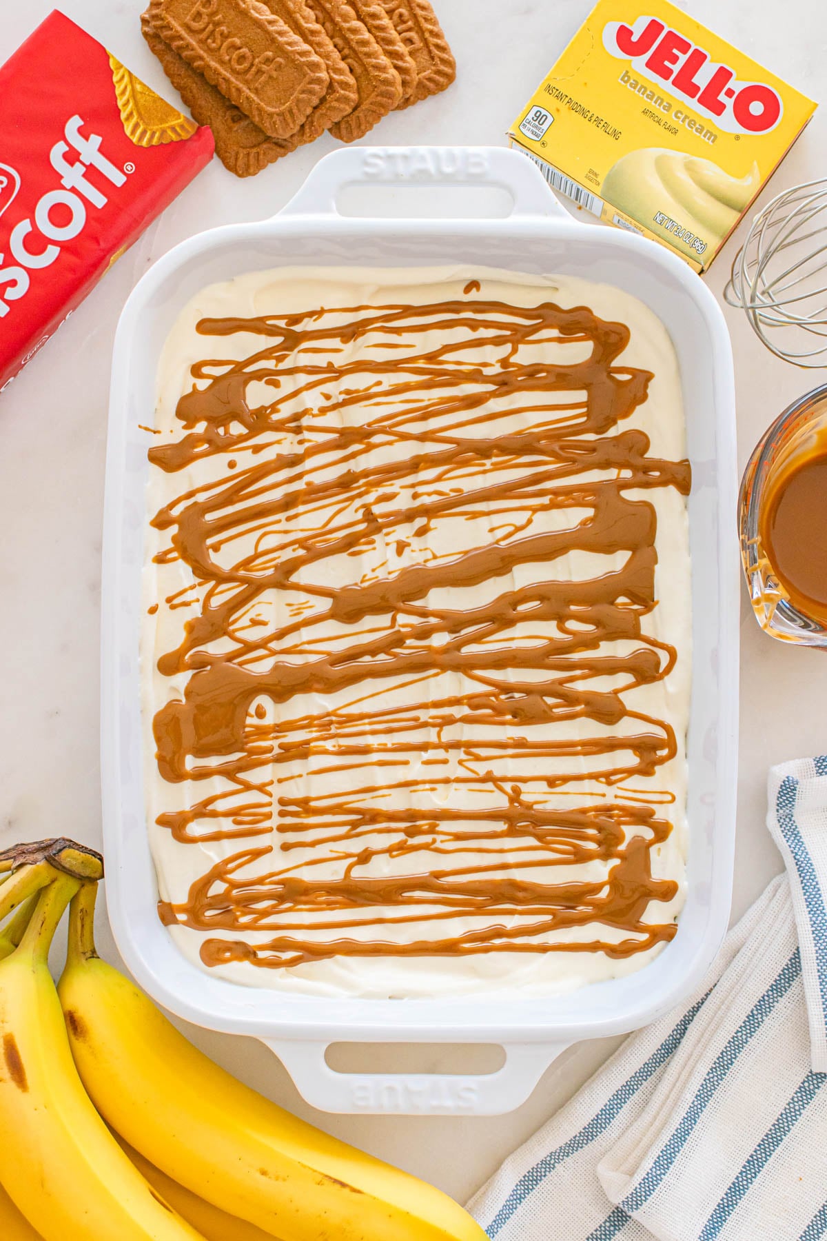 A white baking dish filled with banana cream pudding drizzled with caramel sauce. Ingredients like Biscoff cookies, Jell-O, and bananas surround the dish.
