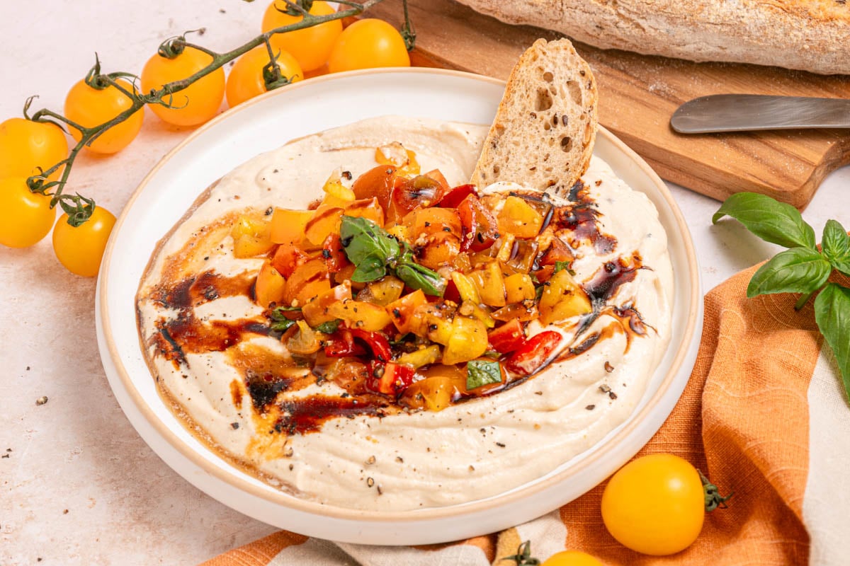 A bowl of dip topped with diced yellow and red tomatoes, chopped herbs, and balsamic glaze, served with a slice of bread. Yellow tomatoes and bread are nearby.