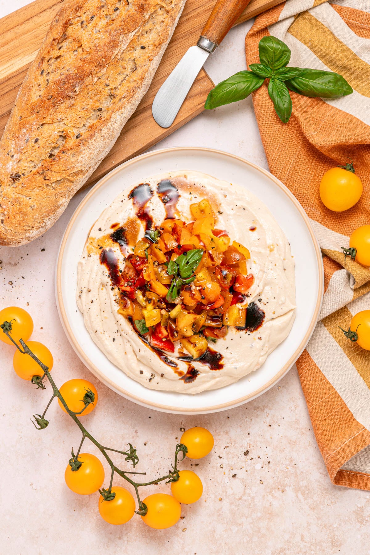 A bowl of dip topped with diced tomatoes, herbs, and balsamic glaze, next to a loaf of bread, yellow tomatoes, and a striped napkin.