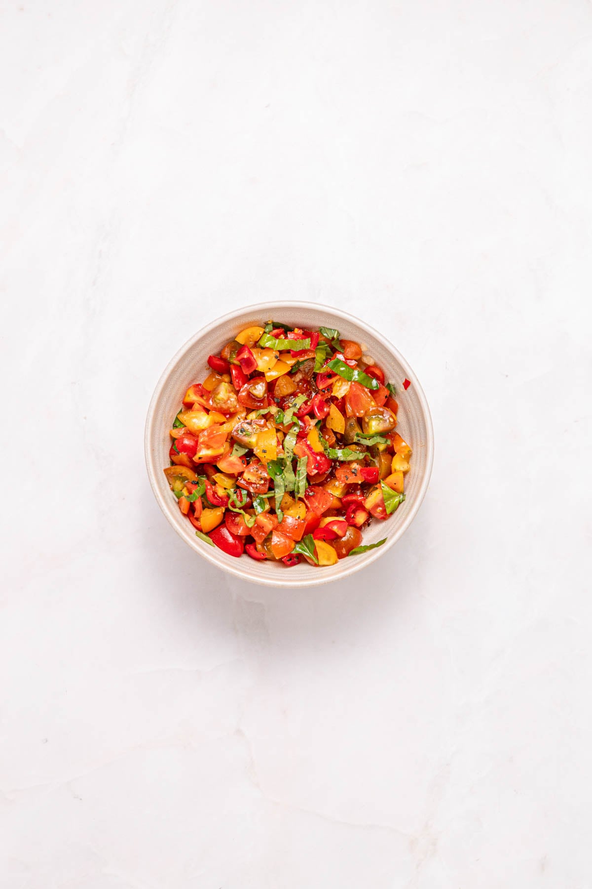 A bowl of colorful chopped tomatoes mixed with herbs is placed on a white marble surface.