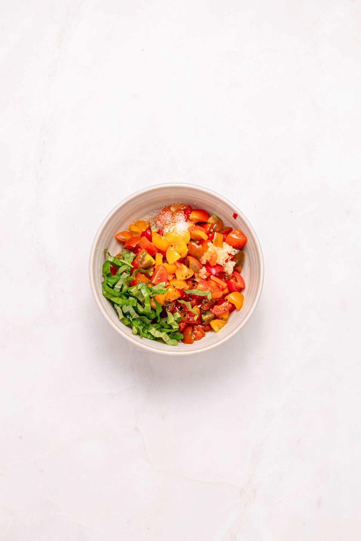 A white bowl containing chopped tomatoes, sliced basil, and seasonings on a light marble surface.
