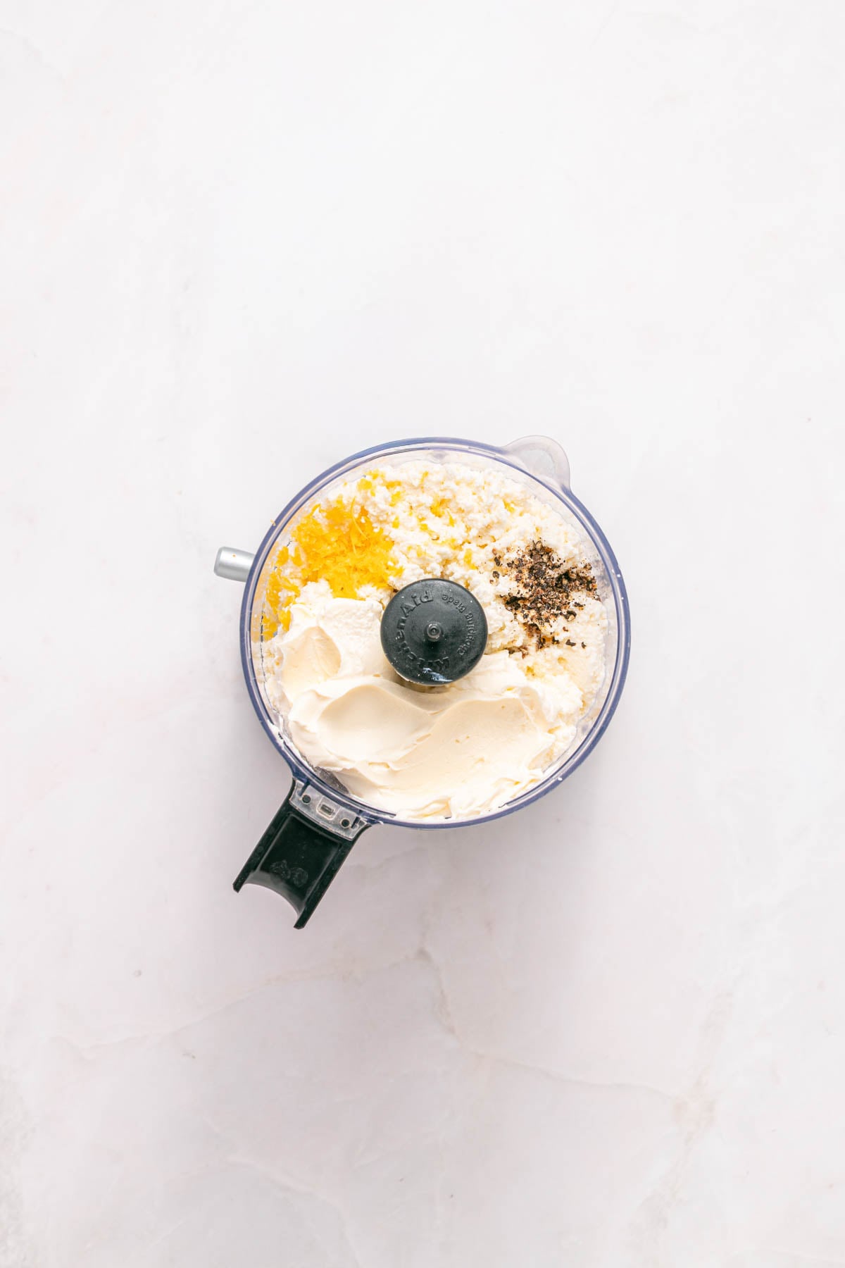 A food processor bowl containing cream cheese, olive oil, lemon zest, and black pepper on a white surface.