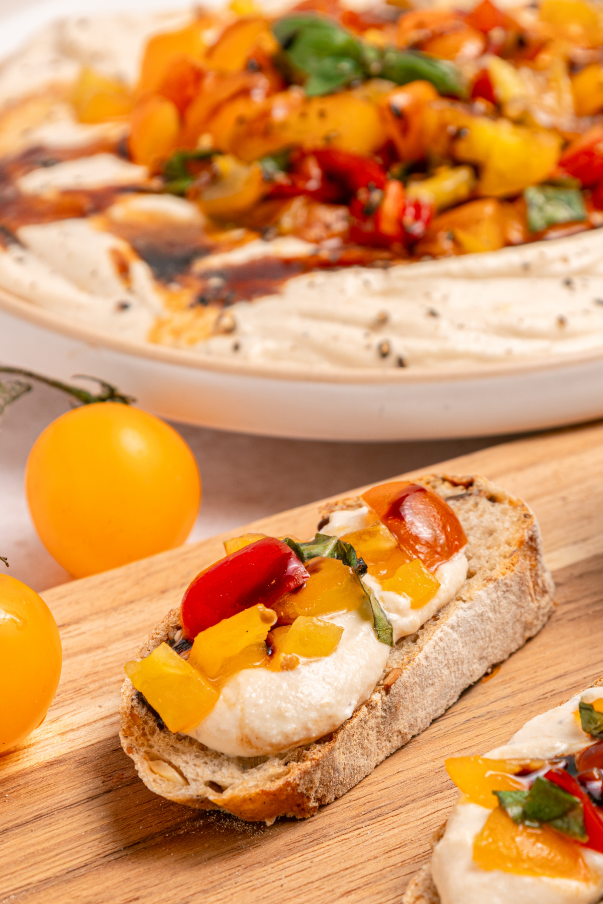 A slice of bread topped with whipped cheese dip, diced yellow and red tomatoes, and basil sits on a wooden board, with a platter of the same ingredients and whole yellow tomatoes in the background.