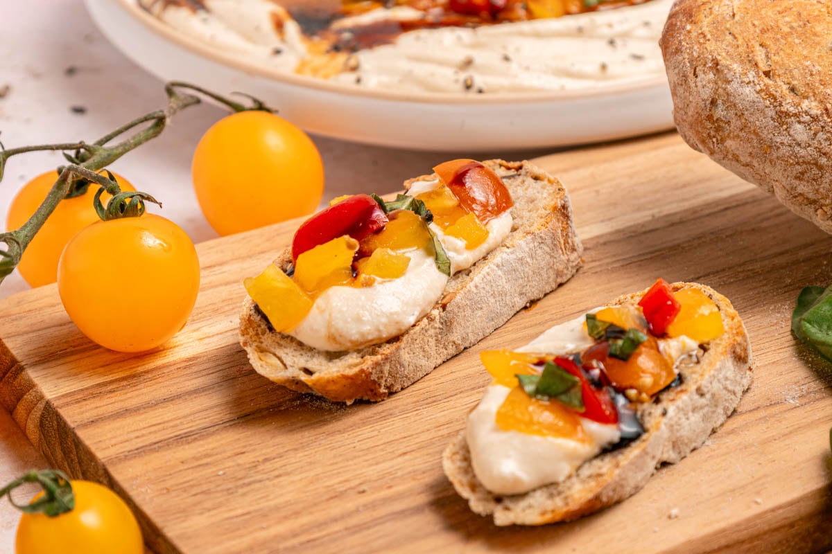 Two slices of bread topped with creamy spread, diced yellow and red tomatoes, and herbs on a wooden board, with yellow tomatoes and bread nearby.