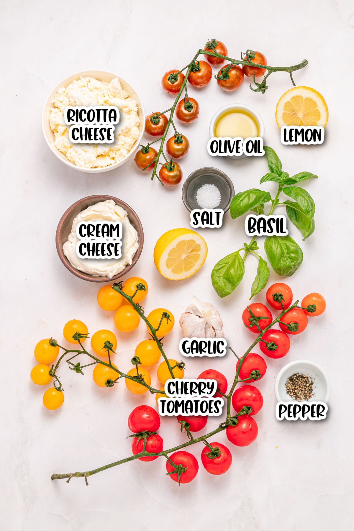 Assorted ingredients on a white surface including ricotta cheese, cream cheese, yellow and red cherry tomatoes, basil, lemon, olive oil, garlic, salt, and pepper.