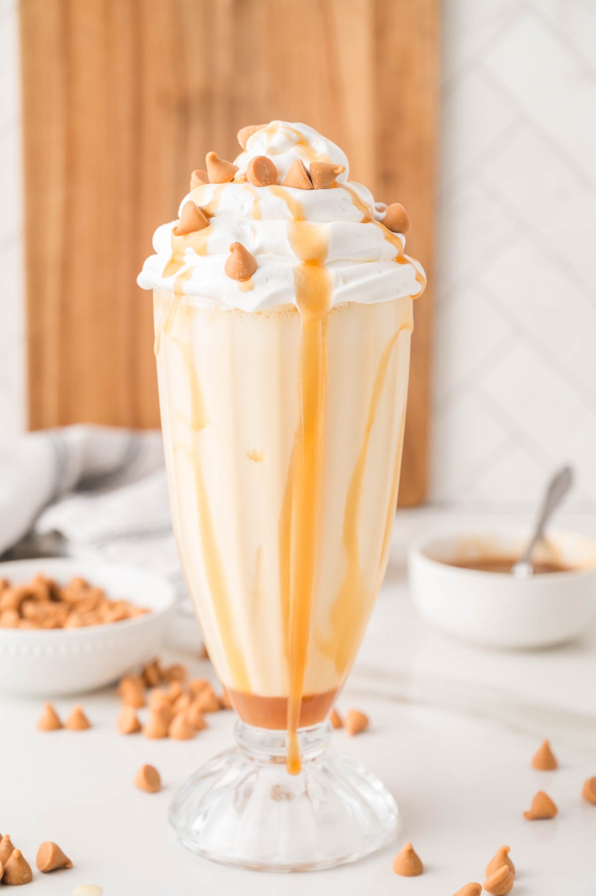 A glass of caramel milkshake topped with whipped cream and butterscotch chips, with caramel sauce dripping down the sides.
