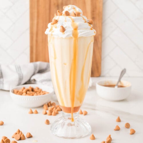 A butterscotch milkshake in a tall glass topped with whipped cream, butterscotch chips, and caramel sauce, with ingredients in bowls nearby on a white countertop.