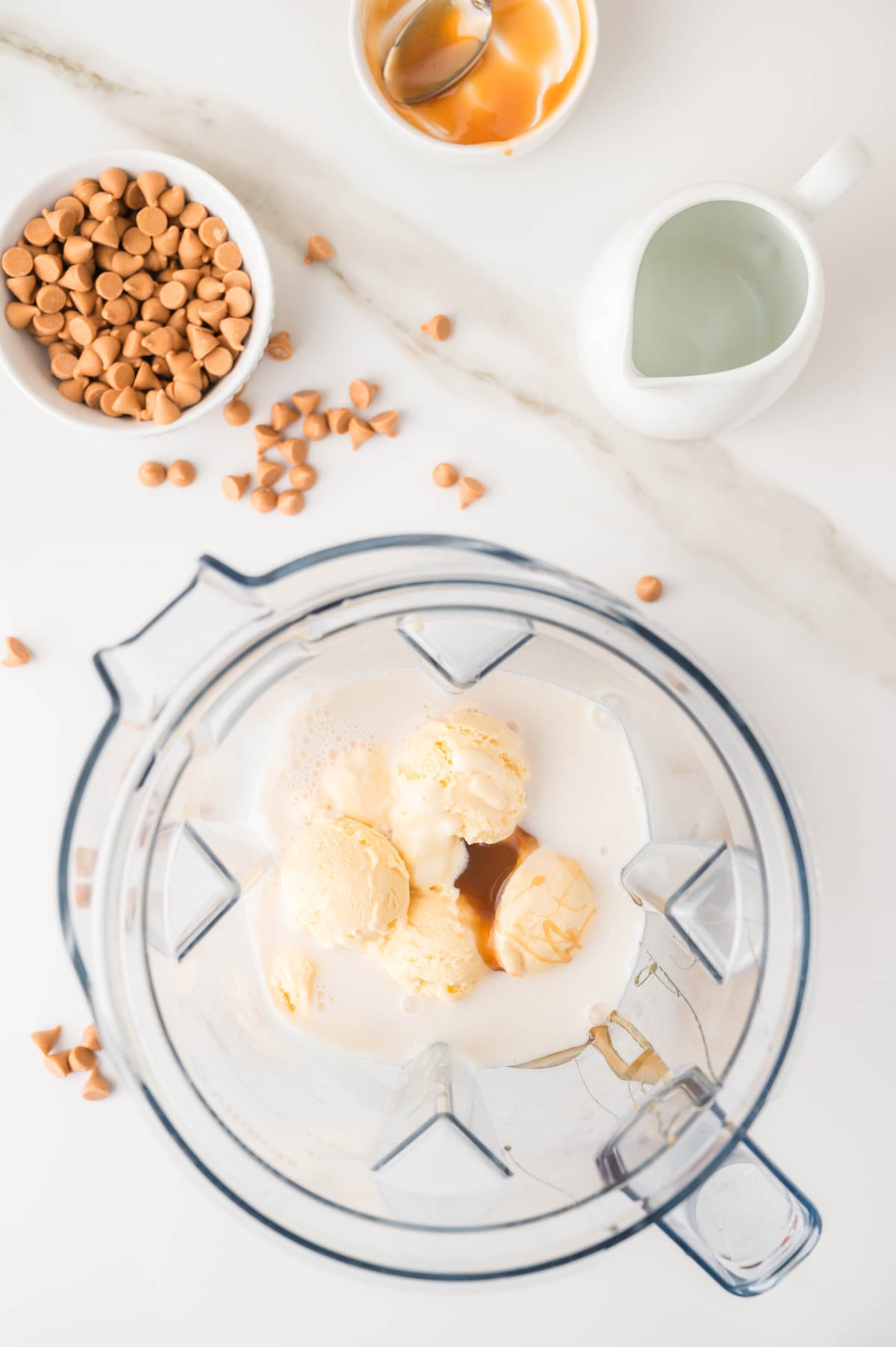 A blender contains vanilla ice cream, milk, and butterscotch sauce. Nearby are a bowl of butterscotch chips, a small pitcher of milk, and an empty bowl with a spoon.