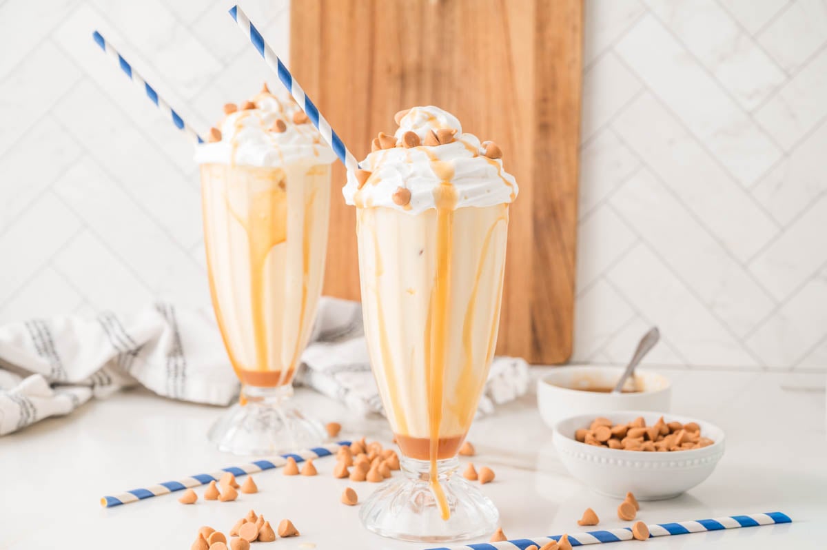 Two tall glasses of a creamy milkshake topped with whipped cream, butterscotch sauce, and butterscotch chips, each with a blue-striped straw, on a white counter.