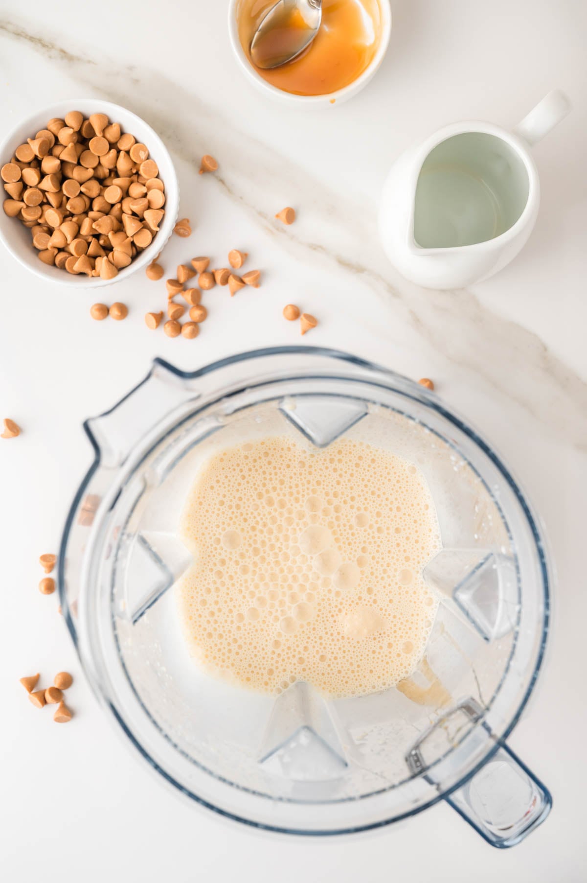 A blender with a frothy mixture inside, surrounded by a bowl of butterscotch chips, a small pitcher of liquid, and a bowl with a spoon and amber liquid on a white countertop.
