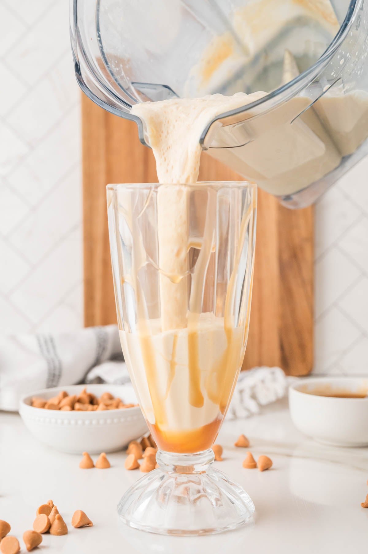 A creamy milkshake is being poured from a blender into a clear glass with caramel sauce drizzled inside, surrounded by butterscotch chips and a white napkin.