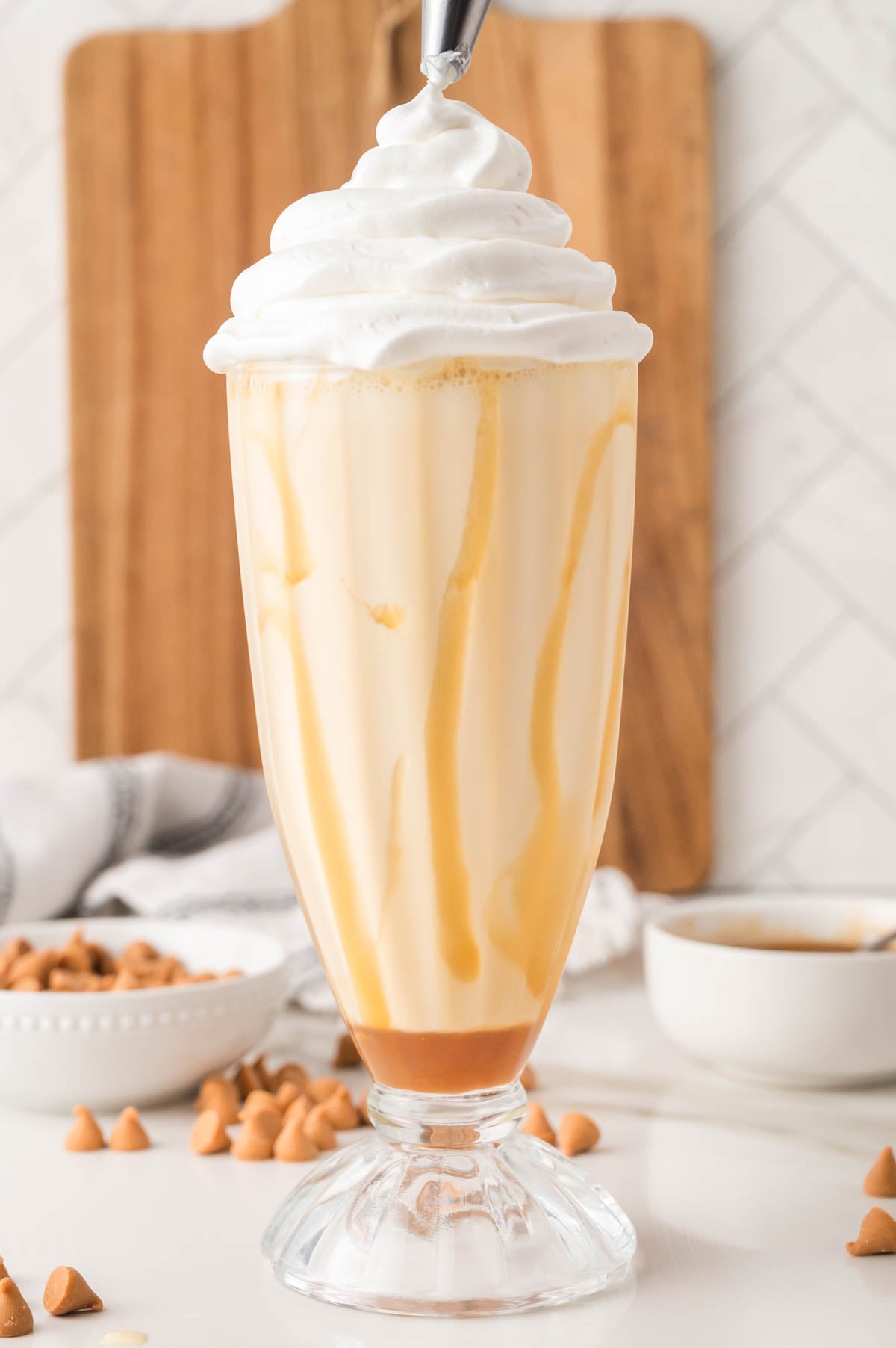 A tall glass filled with a butterscotch milkshake, topped with whipped cream, sits on a white surface with butterscotch chips and a bowl in the background.