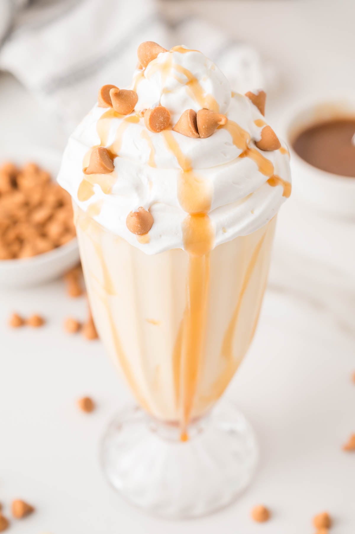A butterscotch milkshake topped with whipped cream, butterscotch drizzle, and butterscotch chips in a clear glass, with scattered chips and a bowl in the background.