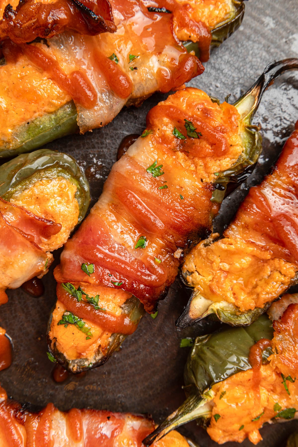 Bacon-wrapped jalapeño peppers stuffed with a creamy orange filling, garnished with chopped herbs, arranged on a dark plate.