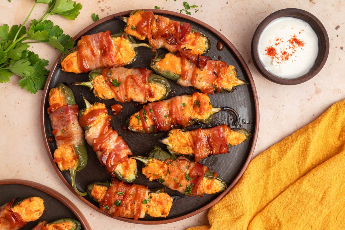 A plate of bacon-wrapped stuffed jalapeños garnished with herbs, accompanied by a bowl of dipping sauce and a yellow napkin on the side.